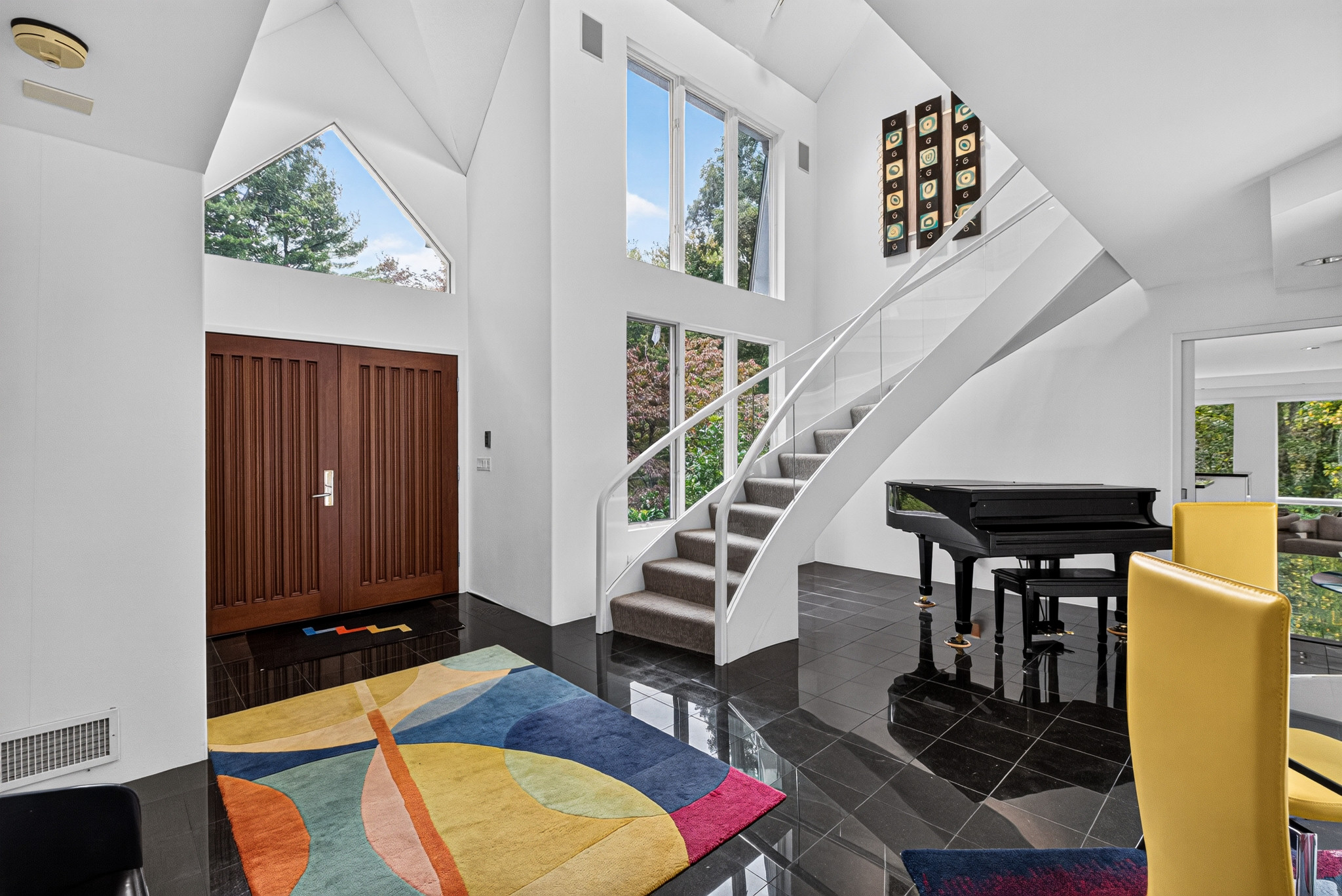 Light-filled foyer with a white staircase, piano, and colorful rug.